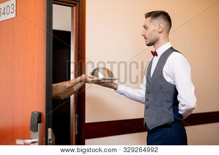 Young serious waiter passing cloche with food to one of hotel guests while standing by open door