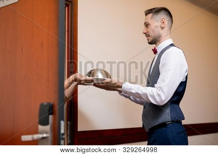Young hospitable waiter in uniform passing cloche with breakfast or lunch to female guest by open door of hotel room