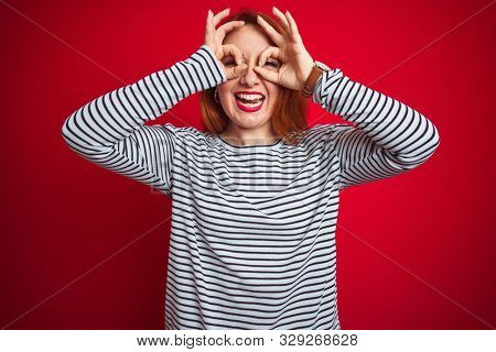 Young redhead woman wearing strapes navy shirt standing over red isolated background doing ok gesture like binoculars sticking tongue out, eyes looking through fingers. Crazy expression.