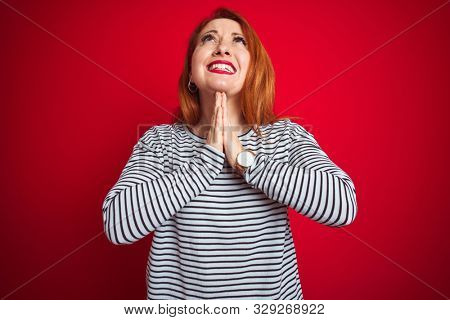 Young redhead woman wearing strapes navy shirt standing over red isolated background begging and praying with hands together with hope expression on face very emotional and worried. Asking