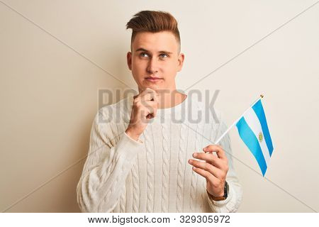 Young handsome man holding Argentinian Argentina flag over isolated white background serious face thinking about question, very confused idea