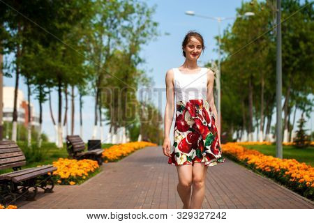Full body portrait of a beautiful young brunette woman walking in flowers dress, summer park outdoors