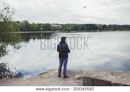 A Man In A Leather Jacket Controls A Drone In The Background Of The Lake
