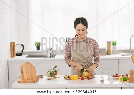 Pretty Young Woman In Apron Smiling While Cuttng Bell Pepper