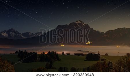Night Starry Sky Over Hohenschwangau Castle. Lake Forggensee In A Mystical Fog. The Allgäu Alps, Bav