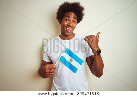 Afro american tourist man holding Argentinian Argentina flag over isolated white background pointing and showing with thumb up to the side with happy face smiling