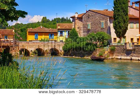 Borghetto Valeggio sul Mincio, Verona, Italy. Italian traditional village with vintage colorful houses above river. Stone bridge among ancient architecture. Boat by bank with stairs. Summer sunny day.