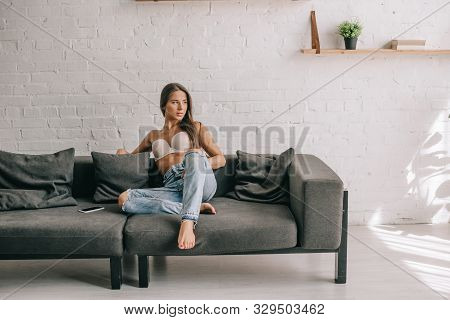 Beautiful Girl In White Bra And Jeans Sitting On Sofa With Smartphone