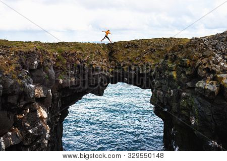 Tourist Jumps Over A Natural Rock Bridge Connecting Basalt Cliffs In Arnarstapi, Iceland, With Atlan