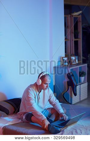 Young Man Wearing Headphones Sitting On The Bed And Working On Laptop Computer Till Night In His Bed