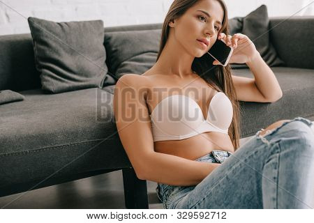 Beautiful Woman In White Bra And Jeans Talking On Smartphone Near Sofa In Living Room