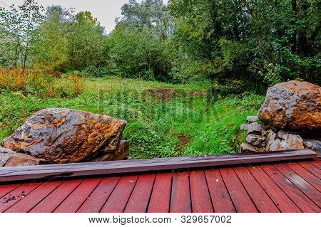 Small Red Wooden Bridge Over A Stream With Two Large Rocks And Trees In The Background, Day At The B