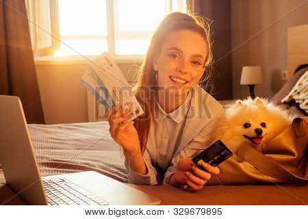 Happy Redhaired Ginger Woman Lying Down In Bed With Her Cute White Fluffy Dog Spitz In Travel Bag Ca