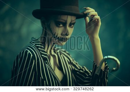 Elegant young woman in a male tailcoat and top-hat with pumpkin skull make-up. Pumpkin queen. Costumes and makeup for Halloween.