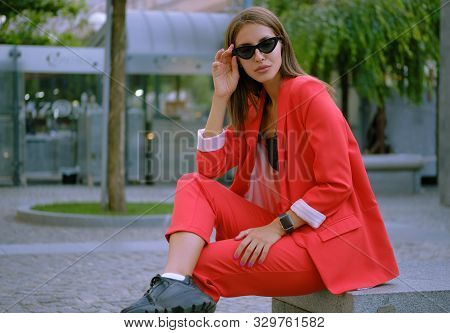 Blonde Girl In A Red Lady-type Pantsuit And Black Top, Watch, Ring, Pendant And Sunglasses Is Posing
