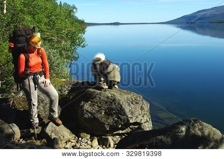 Red-haired Tourist Girl With A Wolfspitz On A Mountain Lake. The Dog Is Standing On A Large Stone On
