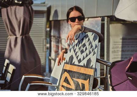 Brunette Girl In Gray Turtleneck, Denim Shorts And Sunglasses Is Posing With Her Wakeboard Satnding 
