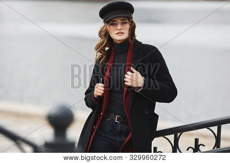 Fashionable Young Woman In A Coat, Modish Hat, And Sunglasses Posing Over An Urban Background. Beaut