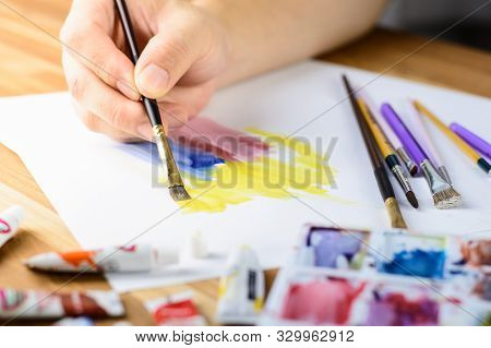 Close Up Of Male Artist's Hand Painting Water Color With Blured Palette With Color Foreground