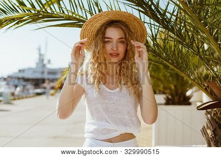 Curly Blonde Holds A Hat In Her Hands And Looks Warily In Front Of Herself On A Background Of Palm T