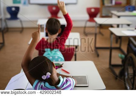 Rear view of african american girl raising her hand while sitting on her desk at elementary school. school and education concept