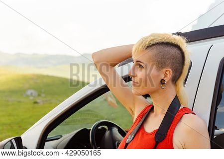 Non-binary Gender Person In A Red T-shirt And Mohawk Hair, With Piercings, Leaning On His Car Thinki