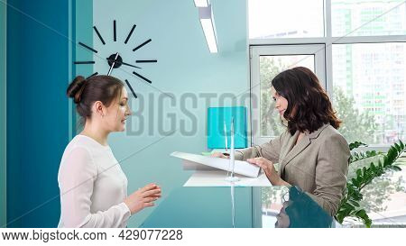 Positive Woman Administrator In White Talks To Client Standing Near Mirror Counter At Reception In C