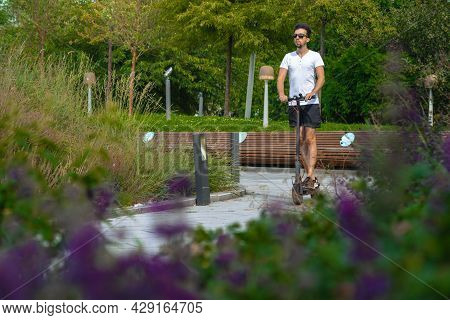 Close Up Of Young Man Riding Black Electric Scooter At Modern Blooming Park In City.