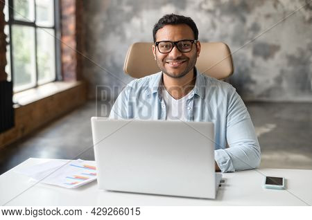 Cheerful Indian Male Student Or Office Worker Is Using Laptop Computer Sitting At The Desk. Friendly