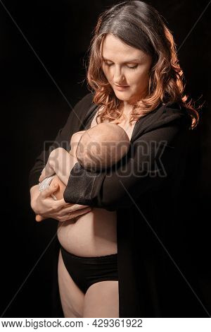 Happy Woman Mother Holds A Sleping Newborn Baby Boy In Her Arms, Black Studio Background. Mom Smiles