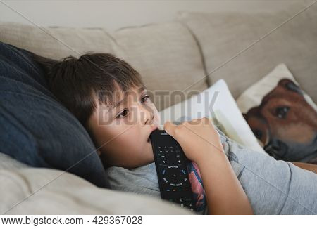 Happy Mixed Race Kid Lying On Sofa Biting Remote Contole While Watching Tv, Child Boy Laying On Couc
