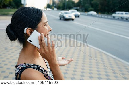Adult Woman With A Hearing Impairment Uses A Hearing Aid In Everyday Life, Talking On Phone In Urban