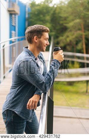 Side Vertical Portrait Of Confident Thoughtful Young Man Standing On Balcony Terrace And Drinking Co