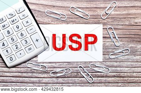 On A Wooden Background, A White Calculator, White Paper Clips And A White Card With The Text Usp Uni