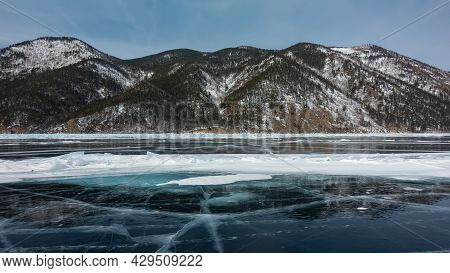Cracks, Snow Spots, Hummocks Are Visible On The Blue Ice Of The Frozen Lake. The Wooded Slopes Of Th
