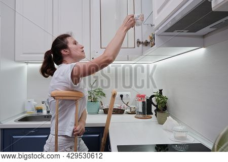 Woman On Crutches Washes The Dishes In The Kitchen. Temporary Limitation Of The Physical Capabilitie
