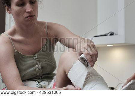 Woman Sitting On A Bed Puts An Elastic Bandage On A Broken Leg. The Leg Is In A Plaster Cast. Home R
