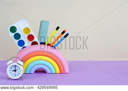 Pencil Case In The Form Of A Bright Rainbow With Paints, Pencils And A Ruler And An Alarm Clock On A