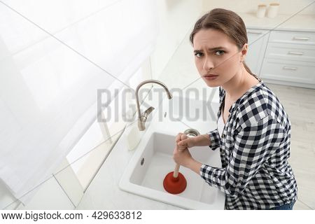 Upset Young Woman Using Plunger To Unclog Sink Drain In Kitchen, Above View