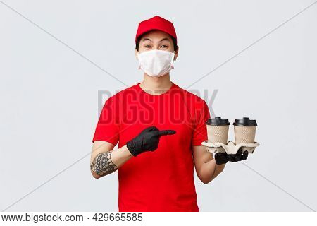 Smiling Asian Delivery Man Red Uniform Cap, Medical Mask And Gloves, Holding Client Order, Pointing 