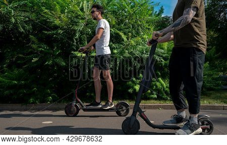Two Friends Driving Electric Scooter In Park And Having Fun At Sunny Day.