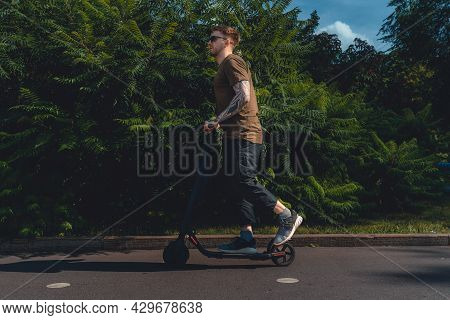 Handsome Young Man On Vacation Having Fun Driving Electric Scooter Outdoors In Modern Park.