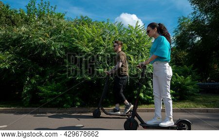 Young Couple Driving Electric Scooters In City Park At Sunny Summer Day