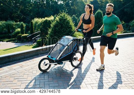 Running Couple With Child In Jogging Stroller In Public Park At Sunrise. Morning Run For A Whole Fam