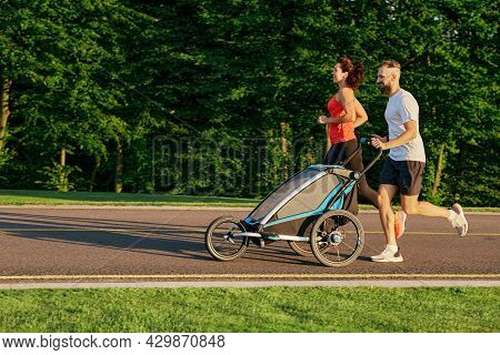Running Couple With Child In Jogging Stroller In Public Park At Sunrise. Morning Run For A Whole Fam