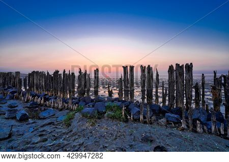 View Of The Wadden Sea During Sunset, At Low Tide. A Colorful Dramatic Sky. Wooden Posts As A Silhou