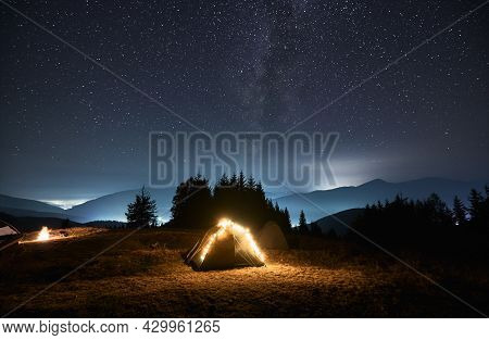 Tourist Tent Lit By Bright Yellow Bulbs On Green Night Lawn Next To Another Tent. Camping On Starry 