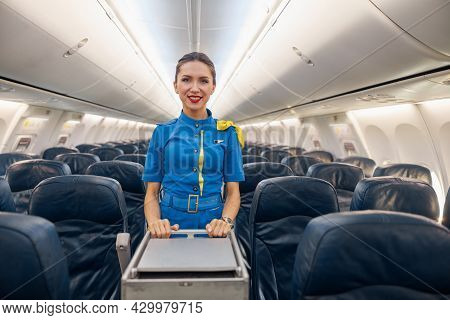Attractive Female Cabin Attendant In Bright Blue Uniform Smiling At Camera While Leading Trolley Car