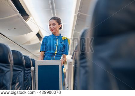 Smiling Female Cabin Attendant Leading Trolley Cart Through Empty Plane Aisle