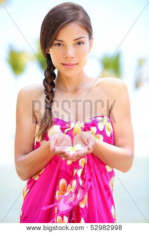 Beautiful serene woman on beach in sarong holding flower in palm of hands. Portrait of pretty graceful mixed race asian young woman on travel vacation on Hawaiian beach, Hawaii, Big Island, USA.